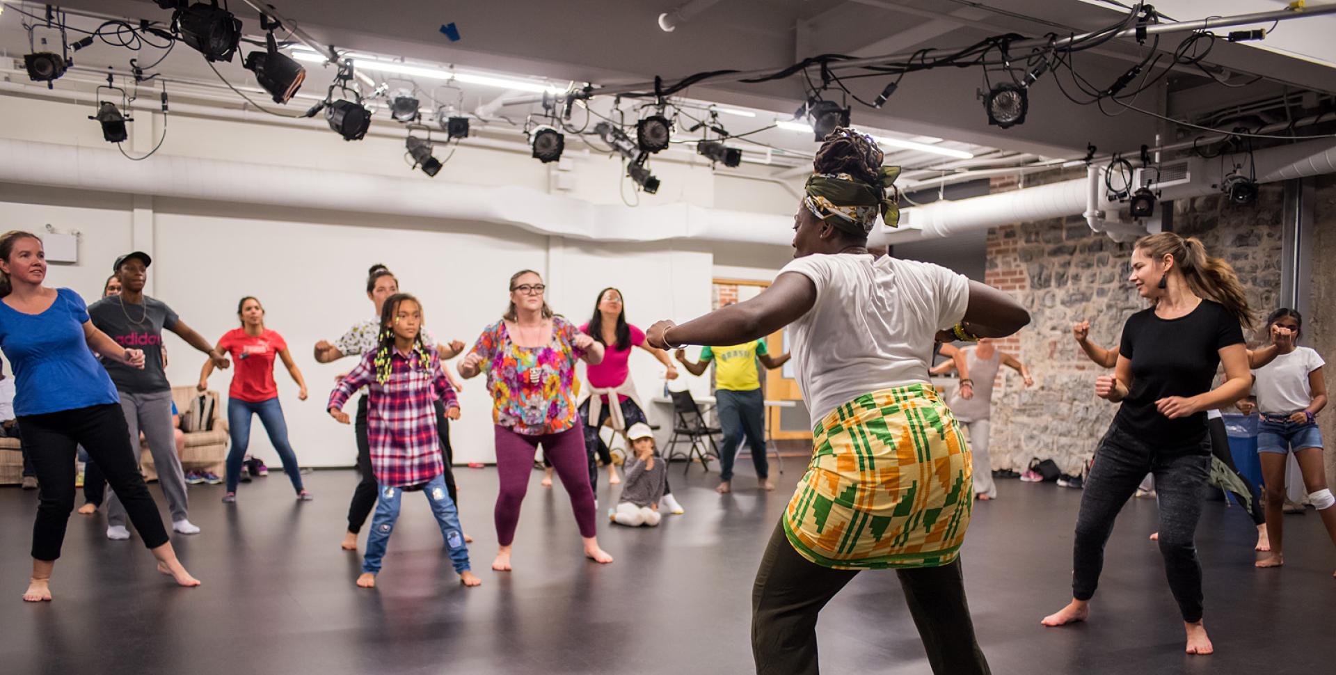 A person with dark skin, a headwrap, and a brightly coloured wrap leads a dance class in the rehearsal hall.