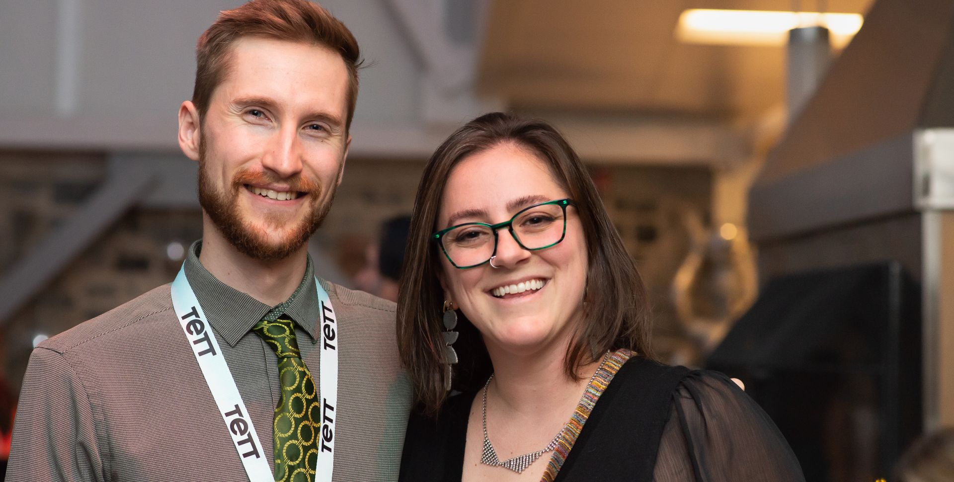 Brandon Elderhorst (Tett Facility Assistant) and Nicole Daniels (Kingston Arts Council's Executive Director) smiling at Tett@10:Reflections. Brandon wears a patterned shirt and Tett lanyard, while Nicole wears glasses and a black top, both looking happy.