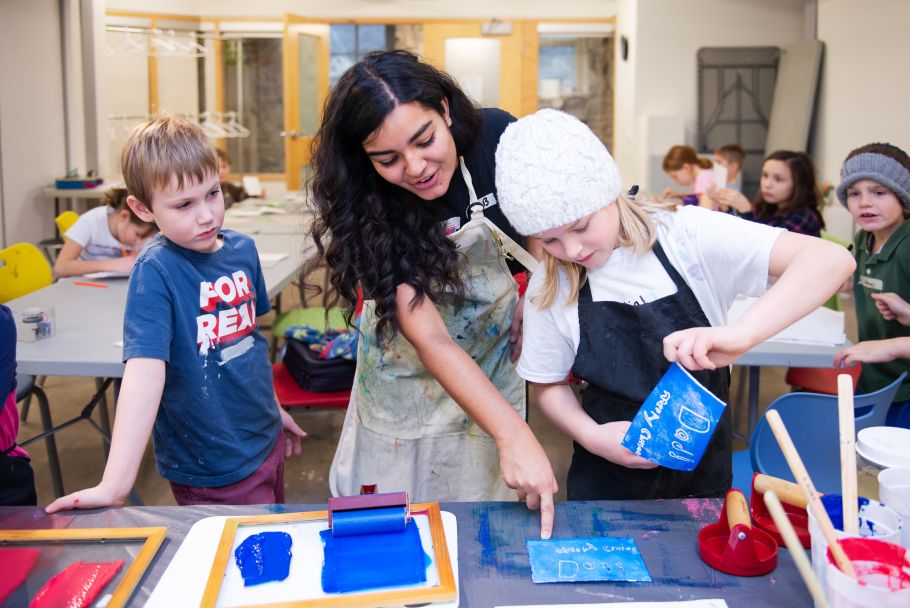 An instructor leads two kids on either side through a printmaking activity.