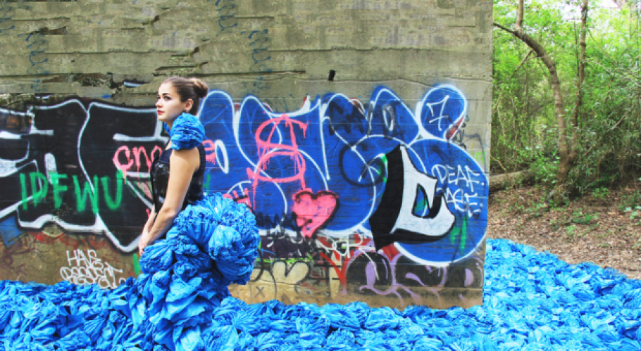 A person with brown hair in a bun wearing an haute couture outfit with blue fabric that cascades in front and behind like water, standing in front of graffiti.