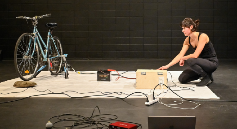 A woman squats down and connects sound cables to a bicycle.