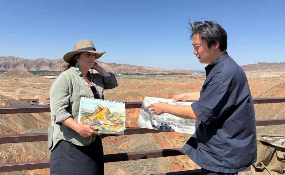 Two people stand on a viewing platform overlooking a rocky landscape, each holding and showing a sketch or drawing of the scenery.