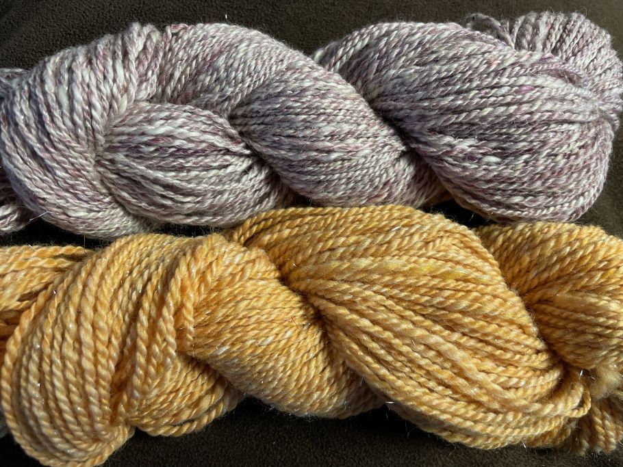 Two skeins of yarn lie side by side on a brown table. The top skein is a blend of lavender and cream, while the bottom is a warm marigold yellow.