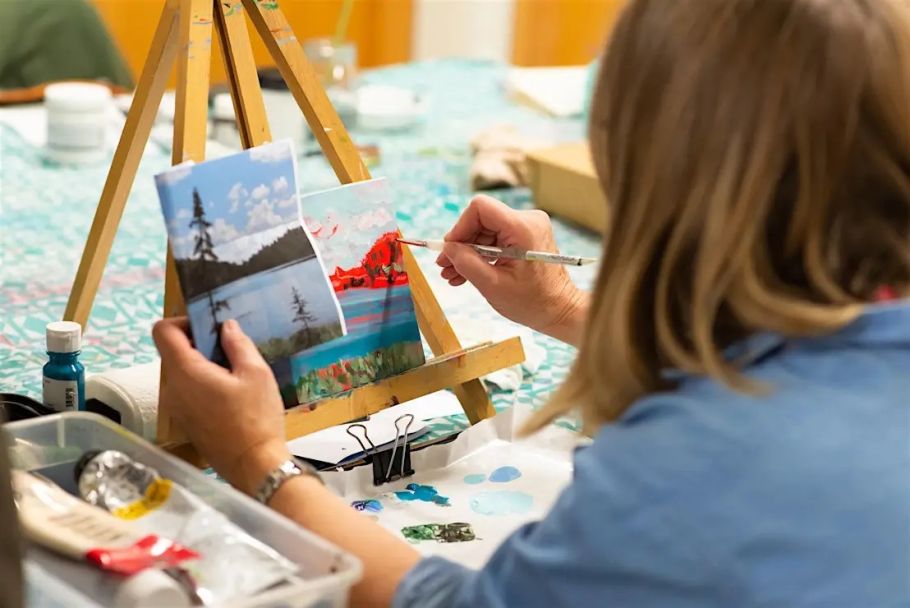 A woman in a blue shirt is painting a landscape on a canvas, using a reference photo clipped beside it.
