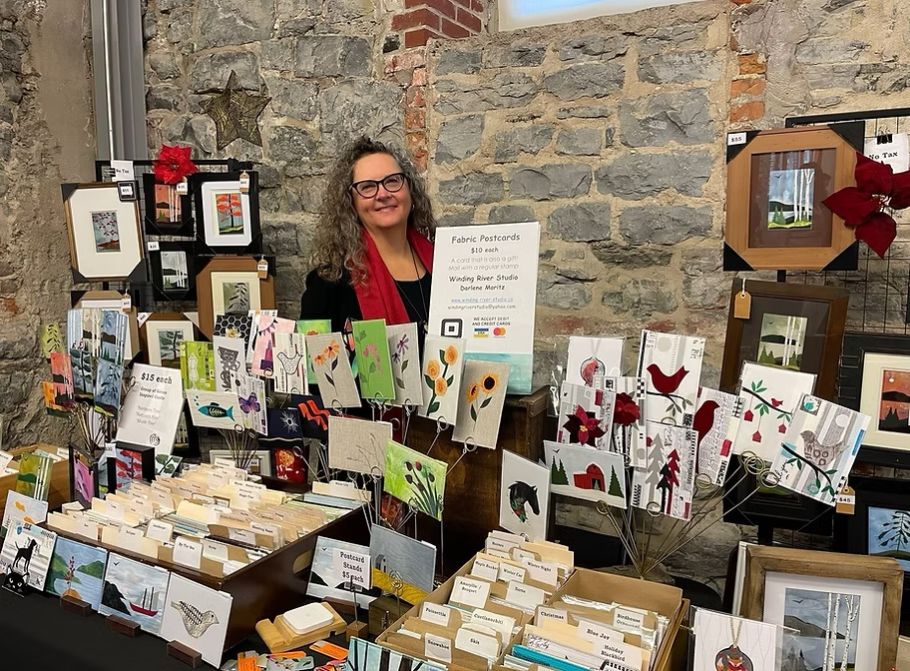 A smiling vendor stands behind a display of colorful fabric postcards and greeting cards at an AWEC craft market. The rustic stone wall adds charm.