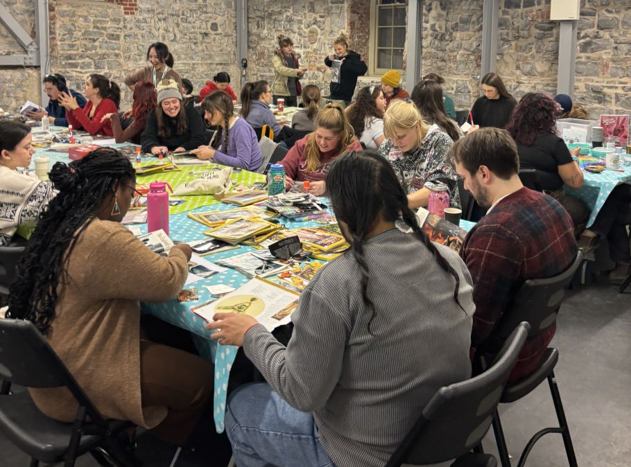 A large group of people sits around tables covered in blue polka-dot cloths, engaged in collage-making with colourful magazines. The atmosphere is lively and focused.
