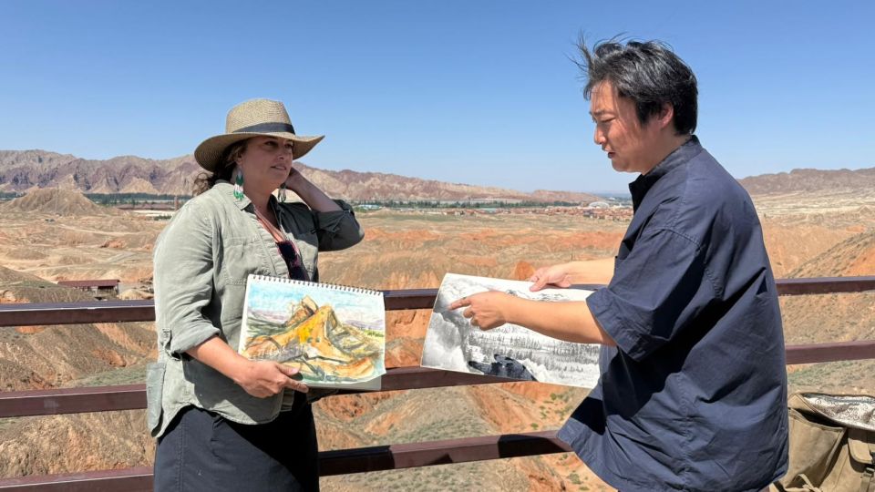 Two people stand on a viewing platform overlooking a rocky landscape, each holding and showing a sketch or drawing of the scenery.
