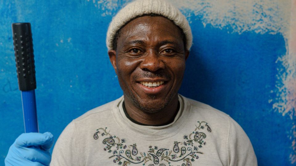 A male-presenting person with Black skin, wearing a white hat and white long sleeved shirt and a blue glove, stands holding a blue broomstick handle. Behind him is a smear of blue paint on a white wall. 