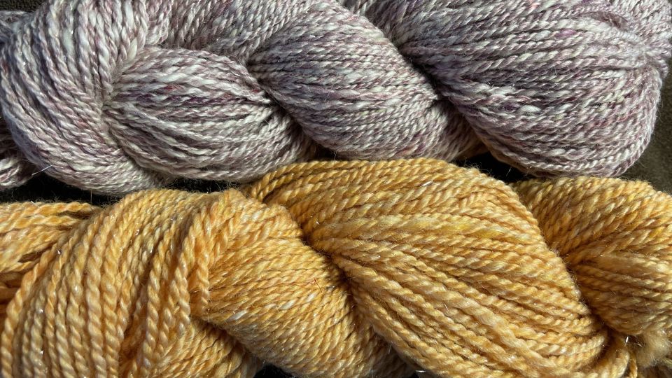 Two skeins of yarn lie side by side on a brown table. The top skein is a blend of lavender and cream, while the bottom is a warm marigold yellow.