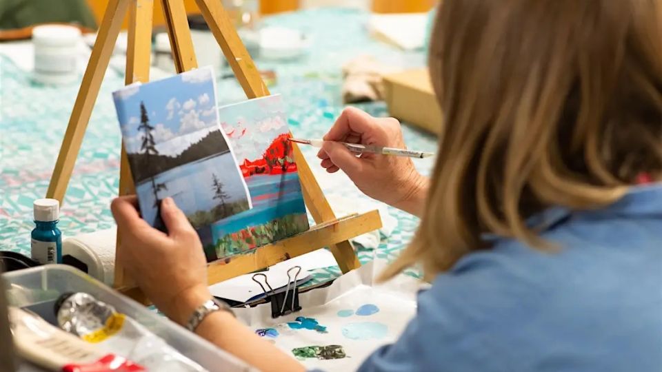 A woman in a blue shirt is painting a landscape on a canvas, using a reference photo clipped beside it.