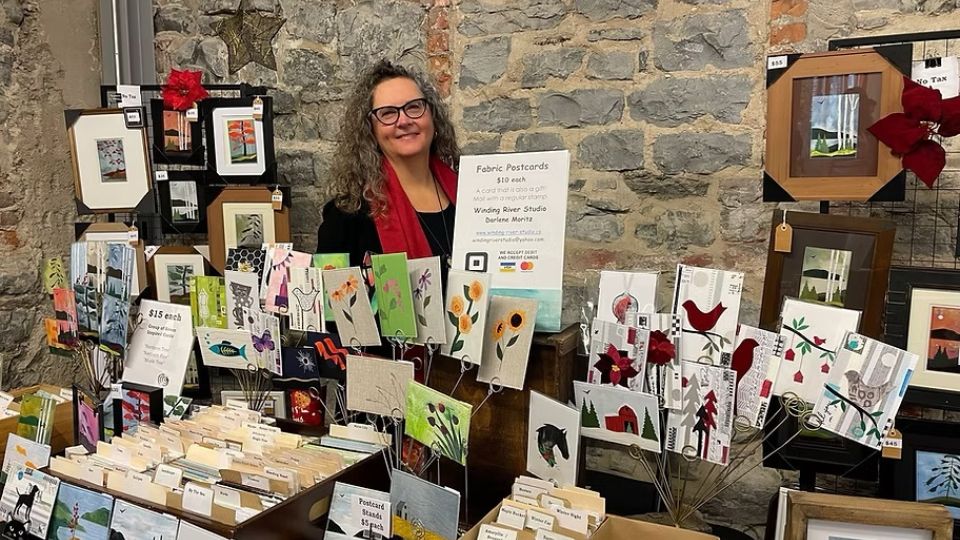 A smiling vendor stands behind a display of colorful fabric postcards and greeting cards at an AWEC craft market. The rustic stone wall adds charm.