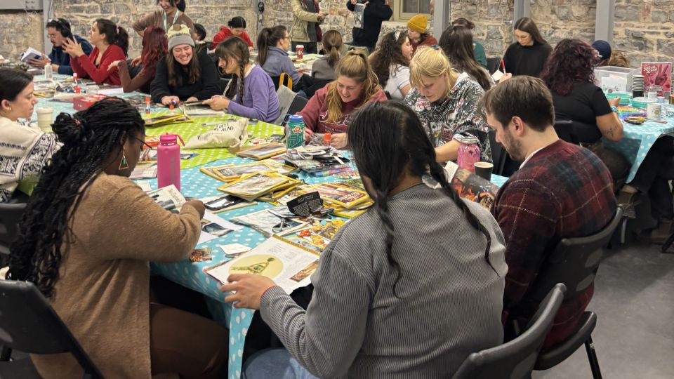 A large group of people sits around tables covered in blue polka-dot cloths, engaged in collage-making with colourful magazines. The atmosphere is lively and focused.