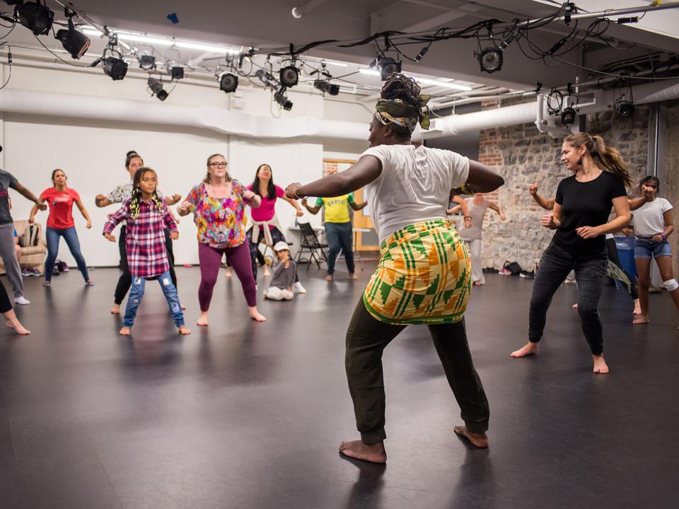 A person with dark skin, a headwrap, and a brightly coloured wrap leads a dance class in the rehearsal hall. 