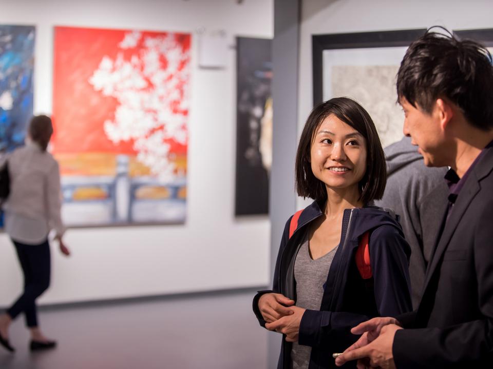 Two smiling people with dark hair and light brown skin smile and talk in front of a gallery space with large canvases. 