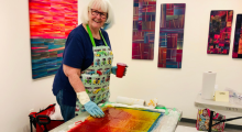 A workshop attendee smiles while dyeing fabric. On the white walls behind her are a series of large colourful fibre works by Bethany Garner. The attendee is a white woman with short white hair with a flash of blue. She wears glasses, a navy blue shirt, jeans and a green apron.